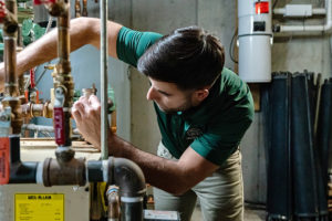 HomeWorks Energy technician in a green shirt inspects a water heater during a heating checkup at a customer's home.